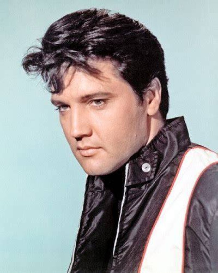 Elvis Presley wears a leather jacket and sits with a guitar.